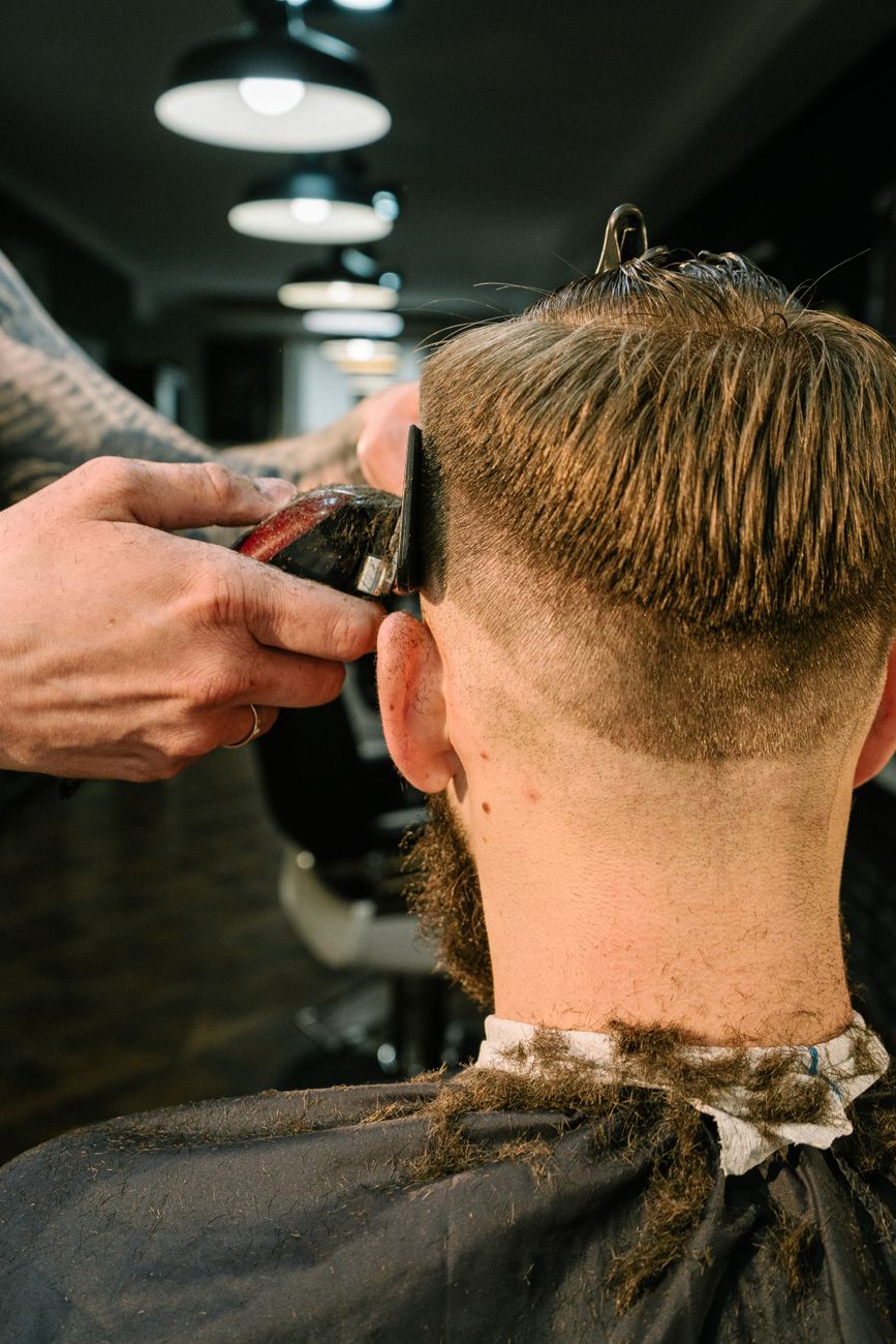Manos de barbero pasando la máquina en la nuca de un cliente sentado, visto desde atrás.