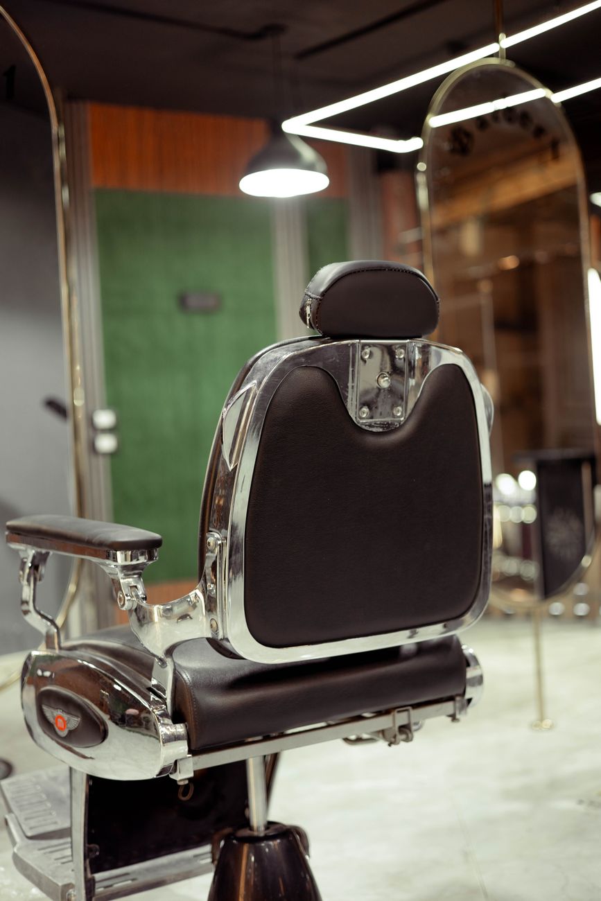 Sillón de barbero de cromo y cuero frente al espejo del local, visto desde atrás.