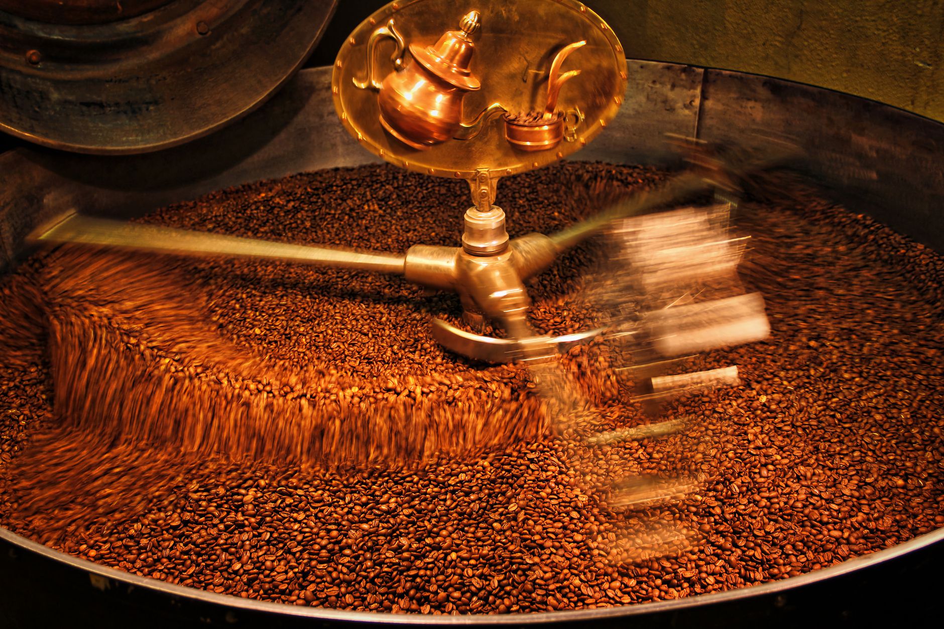 Coffee beans turning in the cooling tray of a drum roaster, the brass arm sweeping through them in a long blur.