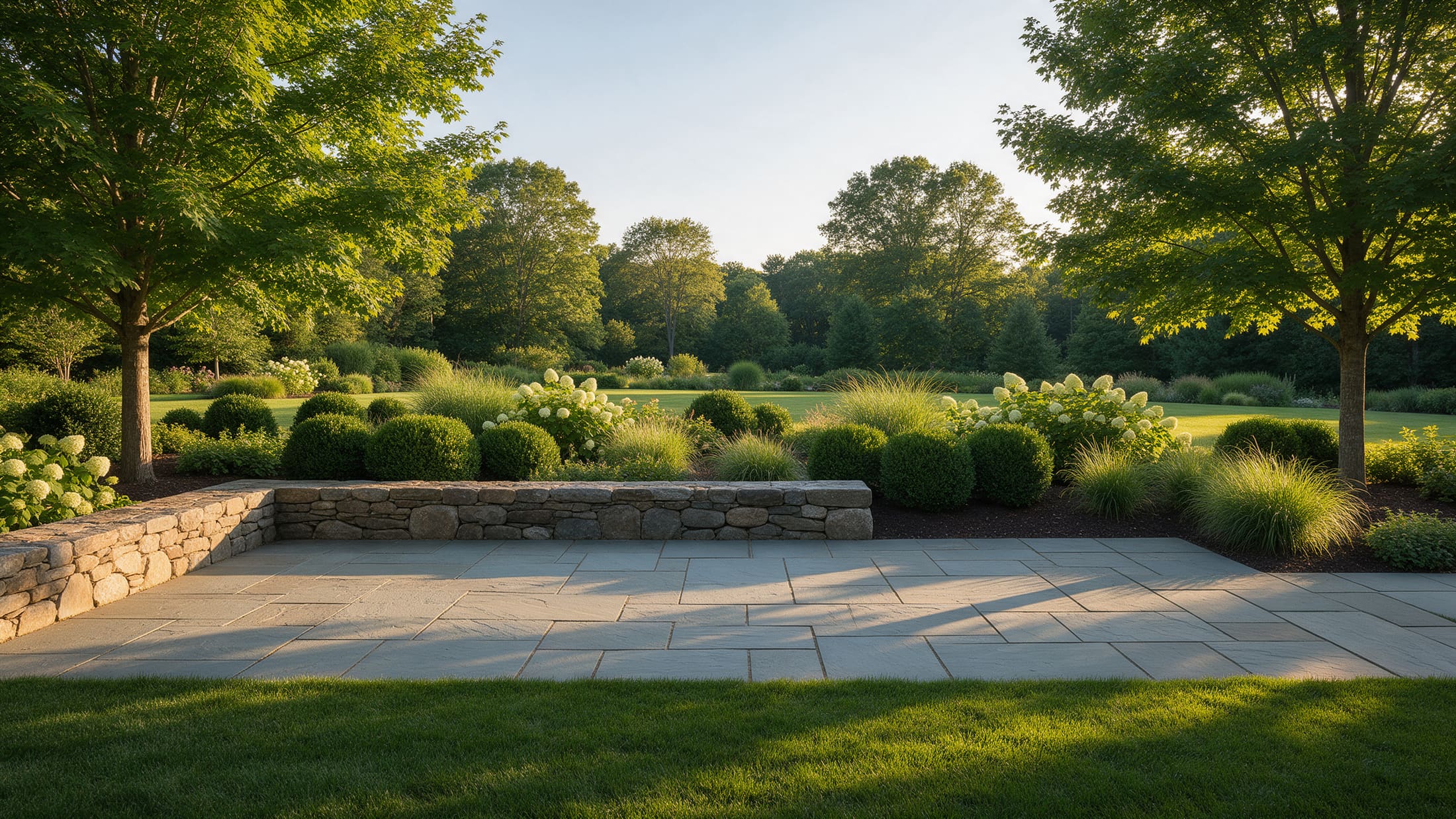 A wide view of a finished garden: a bluestone terrace, a dry-stacked wall, hydrangea and boxwood beds, a lawn and mature trees in low afternoon light.