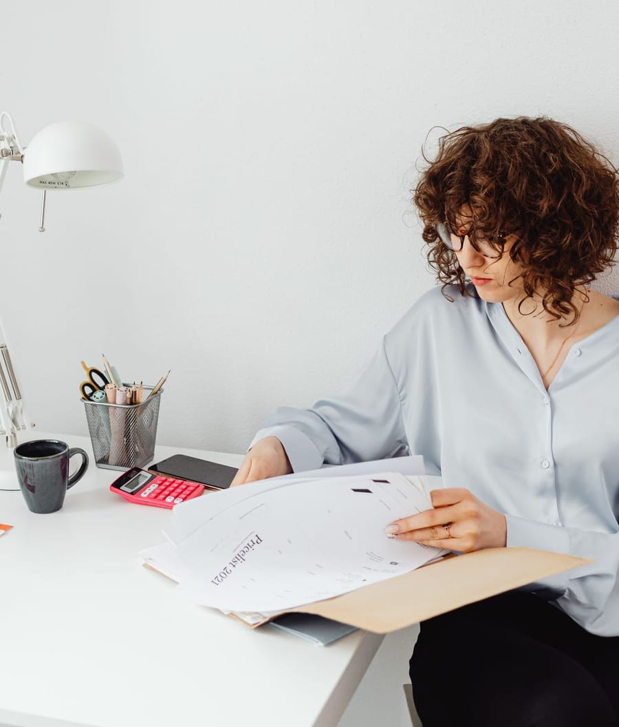 A person at a desk reading through a folder of documents beside a lamp and a cup of coffee.