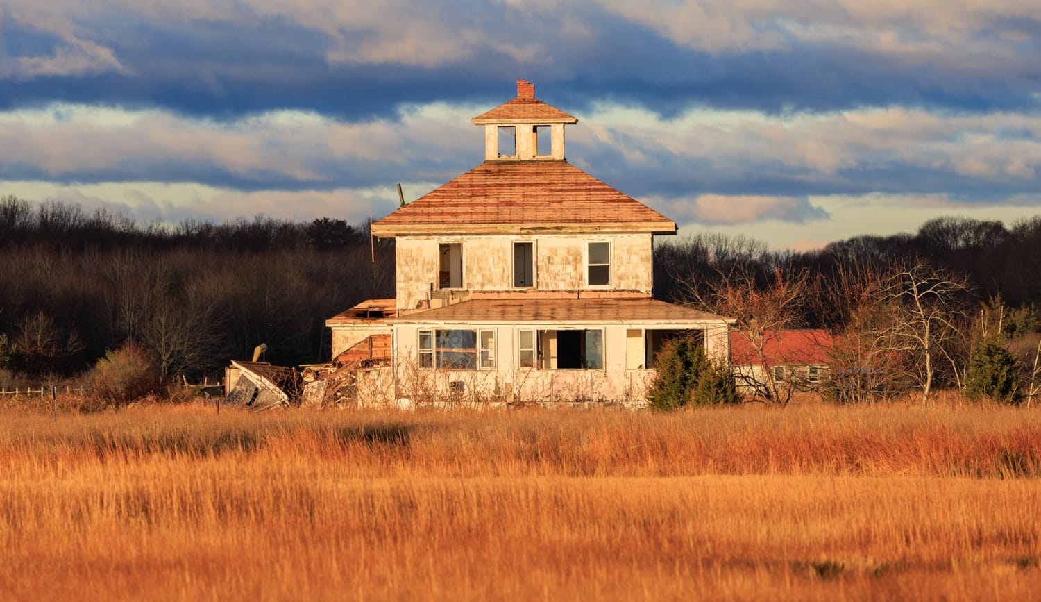 A weathered wooden house standing alone in a salt marsh at low light, with a bank of cloud behind it.