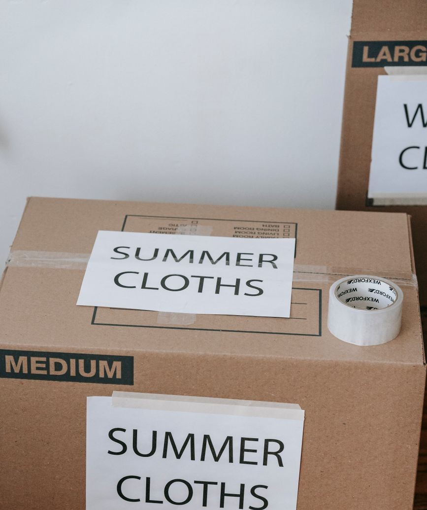 Two cardboard moving boxes with hand-lettered labels reading SUMMER CLOTHS taped to the side, and a roll of packing tape resting on top.