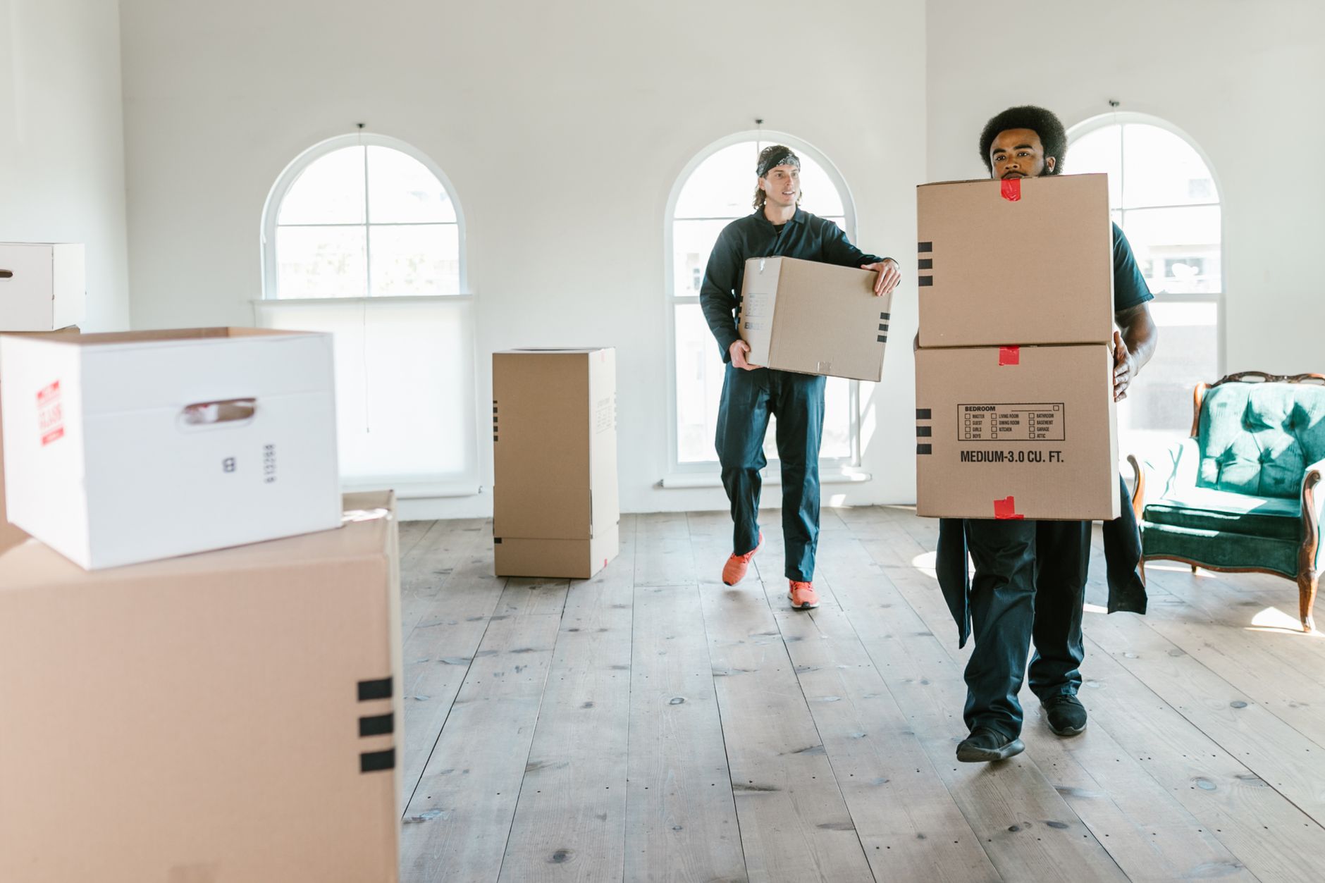 Two movers in navy uniforms carrying labelled cardboard boxes through a bright empty room with tall arched windows and a bare wood floor.