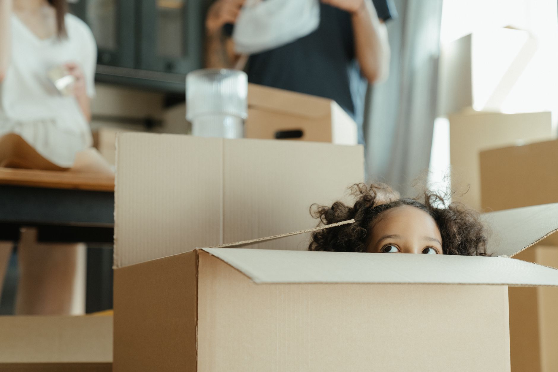 A small child peering over the flaps of an open cardboard box in a half-packed kitchen, with two adults talking in the background.