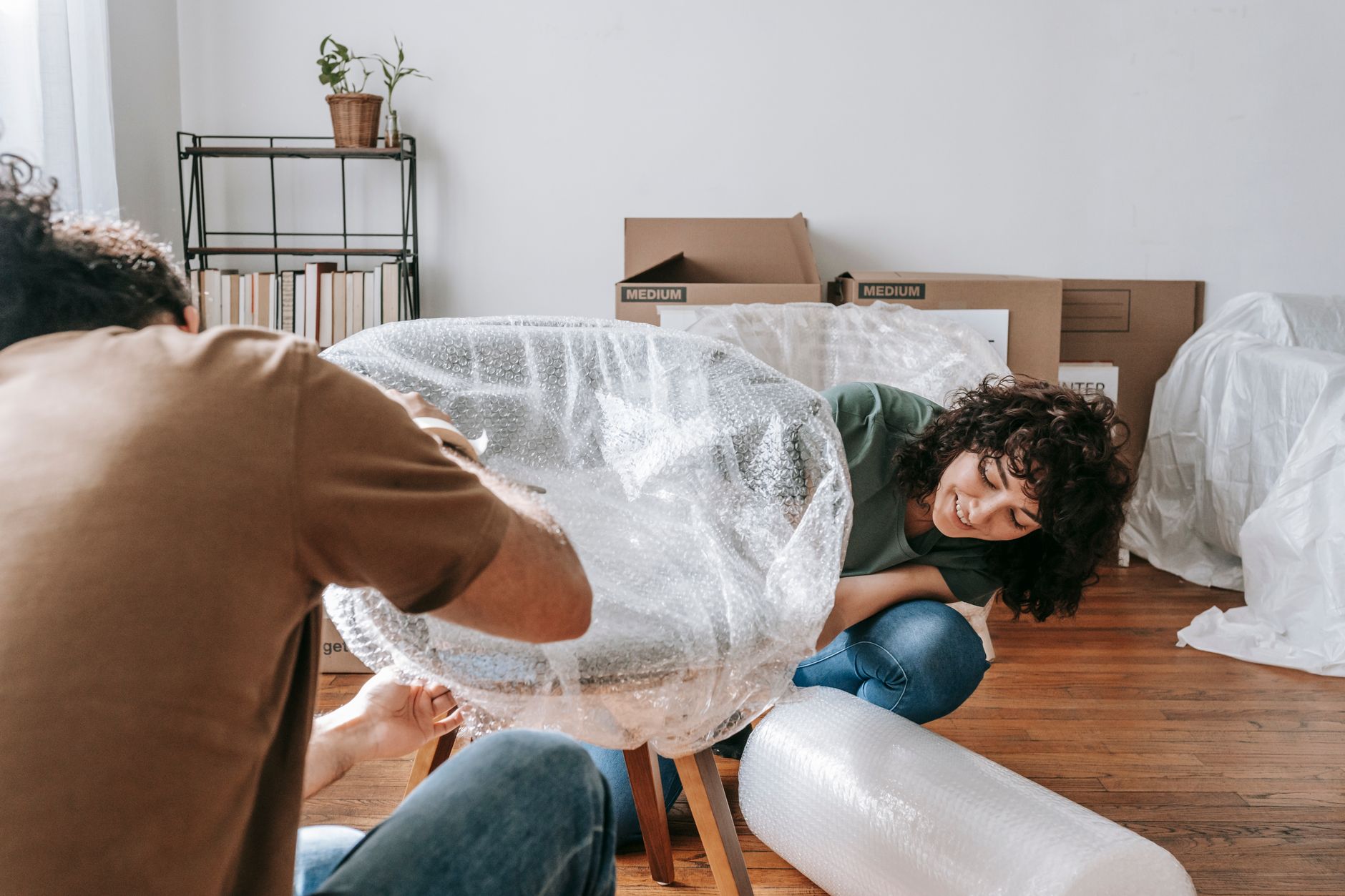Two people wrapping a wooden chair in bubble film on a hardwood floor, with labelled boxes and a covered armchair behind them.