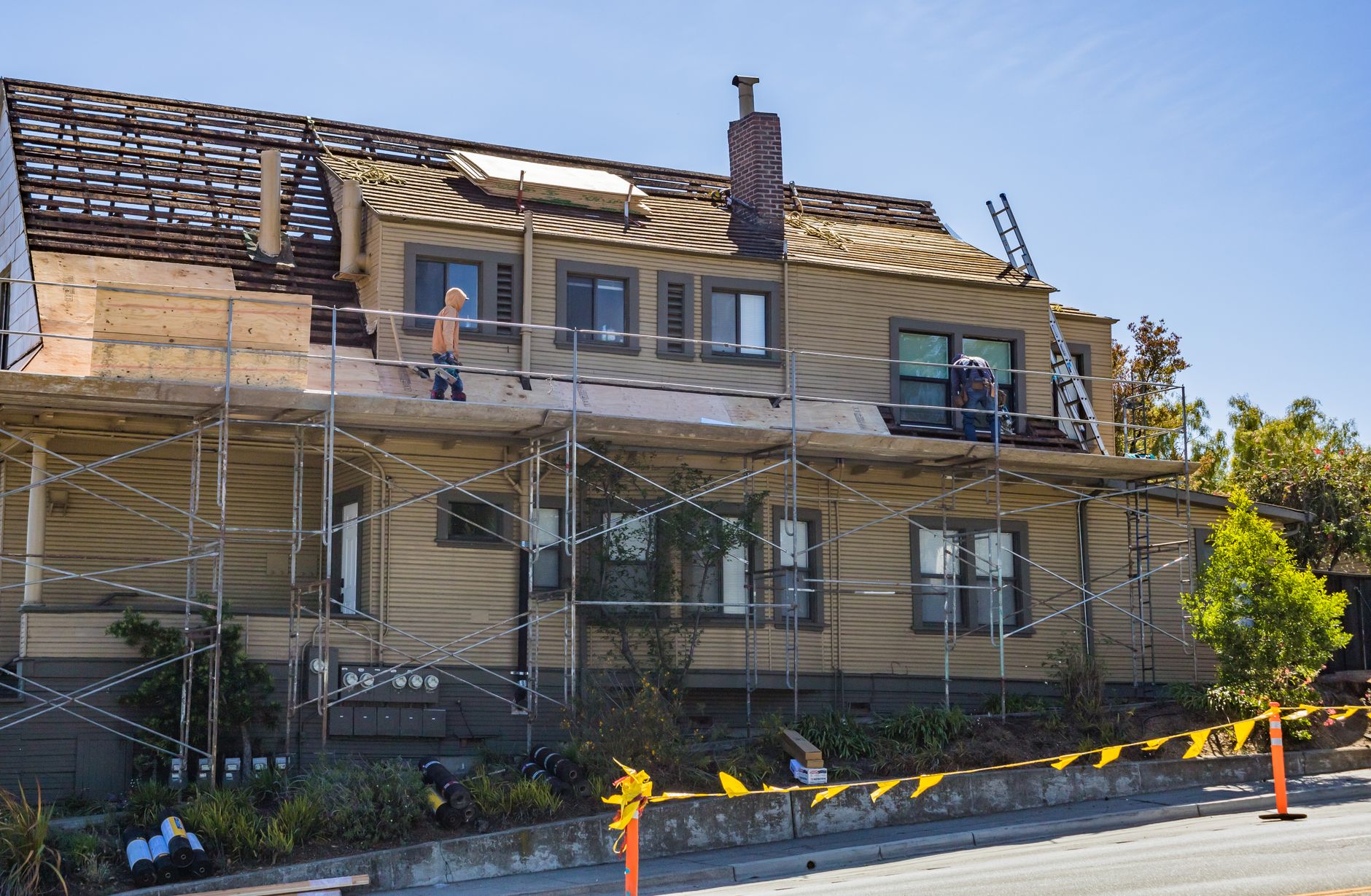 A house stripped back to bare roof boards with scaffolding along the front, plywood stacked on the platform and two workers on the deck.