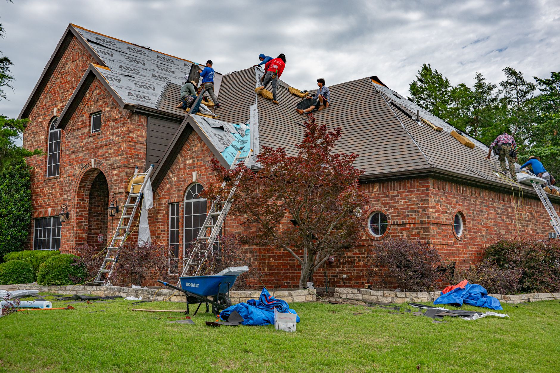 A six-person crew stripping the old shingles off a brick house, with underlayment rolls half unrolled, two ladders up and tarps spread on the lawn below.