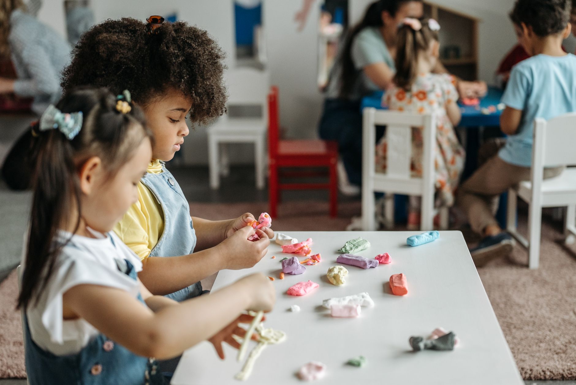 Two young girls shaping coloured clay at a low table in a classroom, with a teacher and three other children working at a second table behind them.