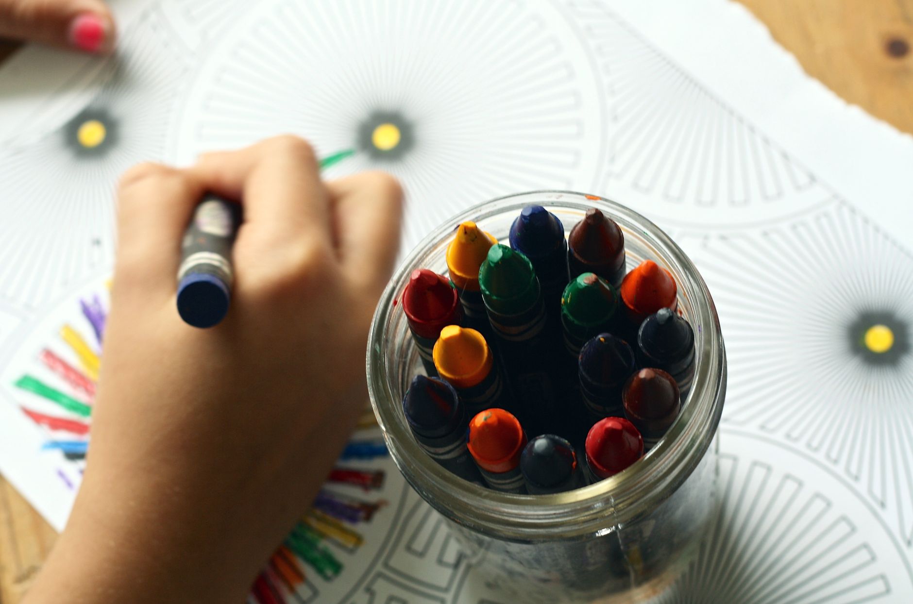 A child's hand drawing on paper beside a glass jar packed with well-used crayons seen from above.