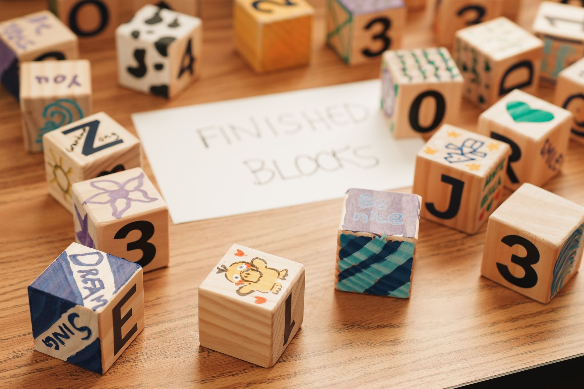 Wooden alphabet and number blocks scattered on a table, each one decorated by hand with drawings and words, beside a card reading Finished Blocks.