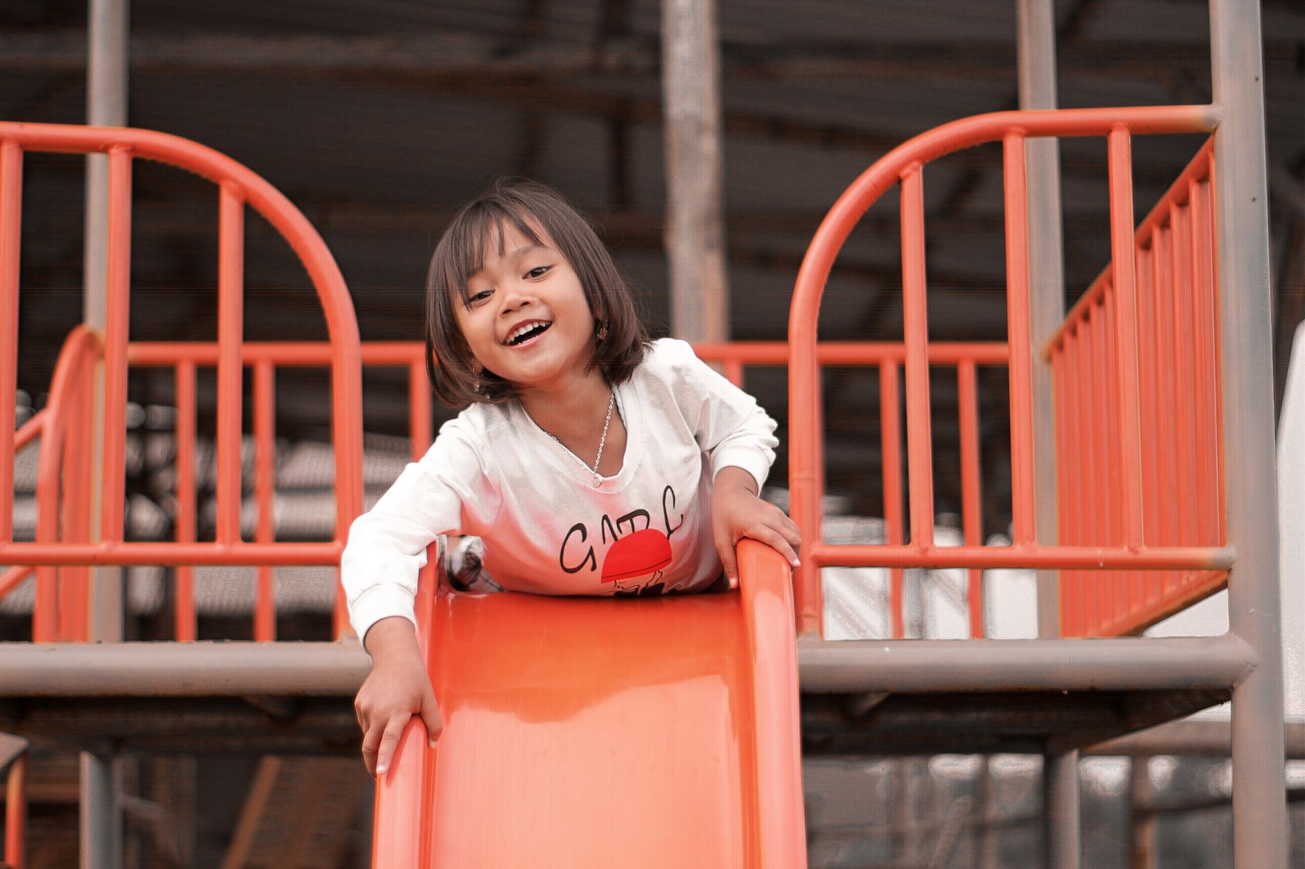 A child in a white long-sleeved top going down a bright orange slide in an outdoor playground.