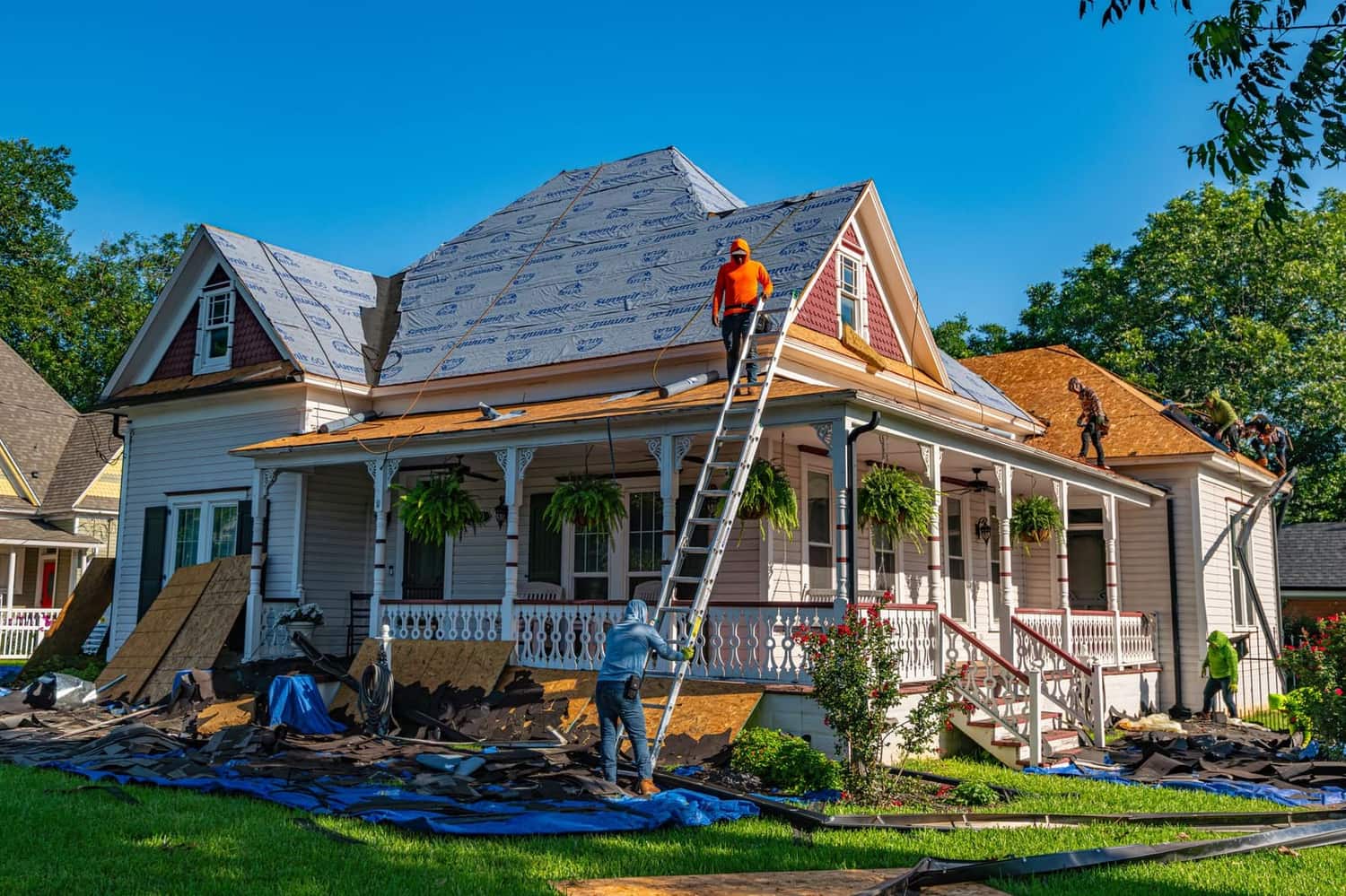 A crew re-roofing a white Victorian house, with tarps and stripped shingles laid out across the front lawn.