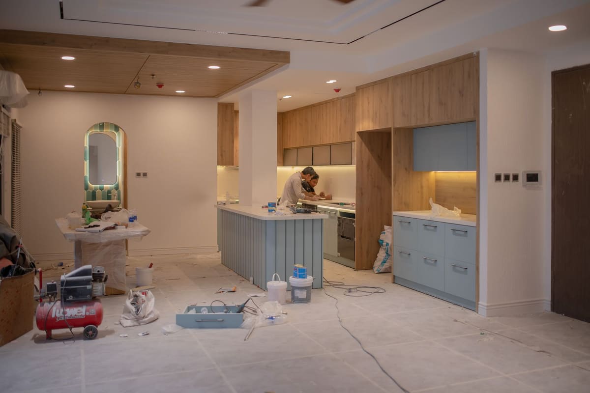 Two carpenters working at a half-installed kitchen: cabinet boxes set, floors papered over, a compressor and buckets on the ground.