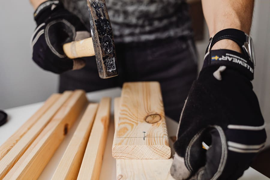 Gloved hands holding a hammer over a pencil-marked pine board with a nail started at the line.