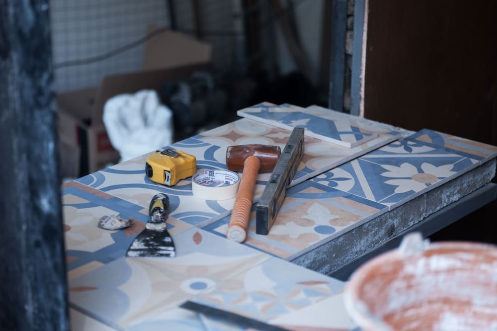 A tape measure, rubber mallet, spirit level and putty knife resting on patterned floor tile laid out on a bench.